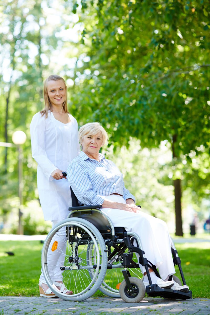 Pretty nurse walking with senior patient in a wheelchair in park